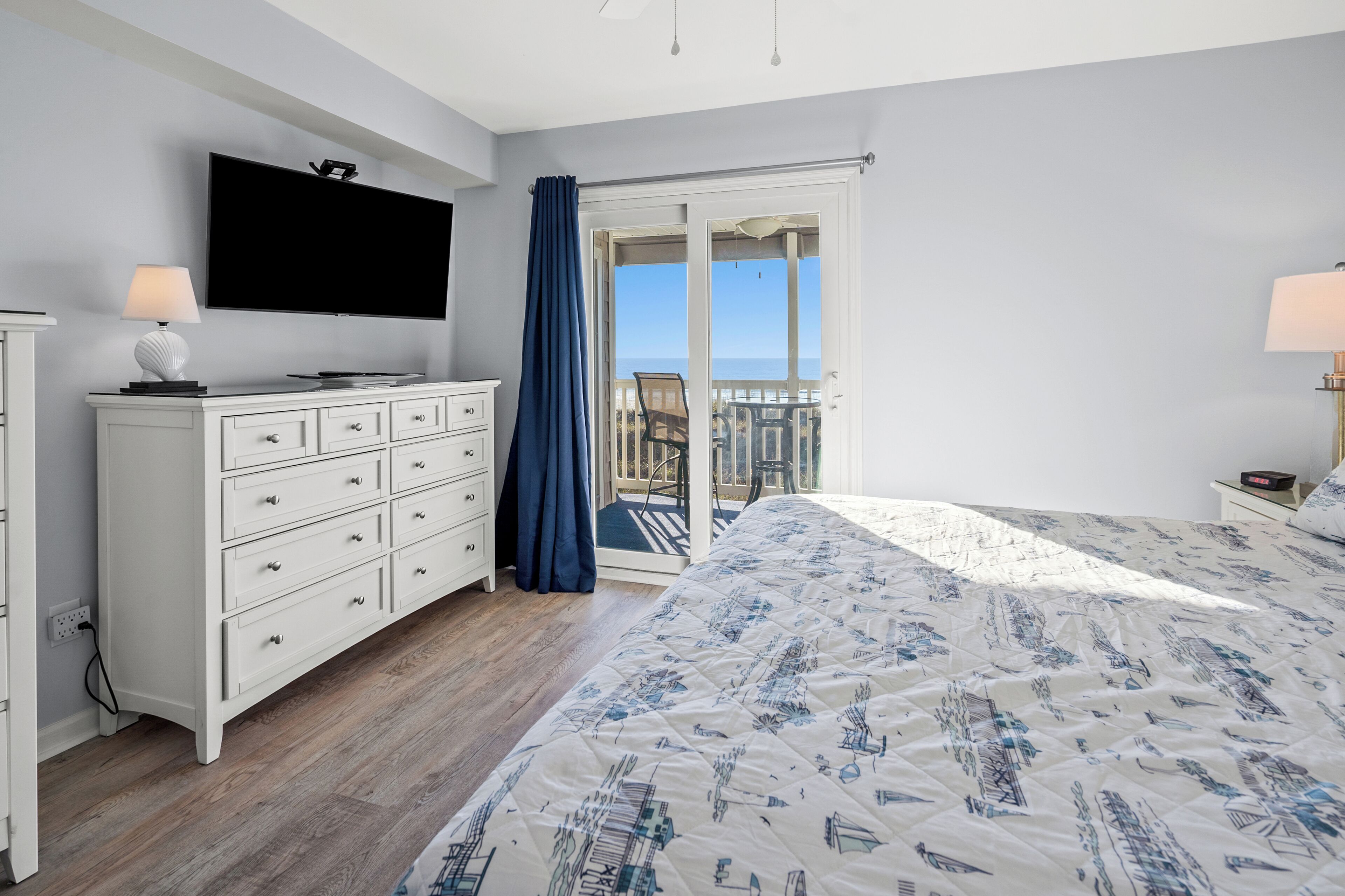 A bedroom with a large bed, a TV mounted on the wall, a dresser, and a balcony with a view of the sea.