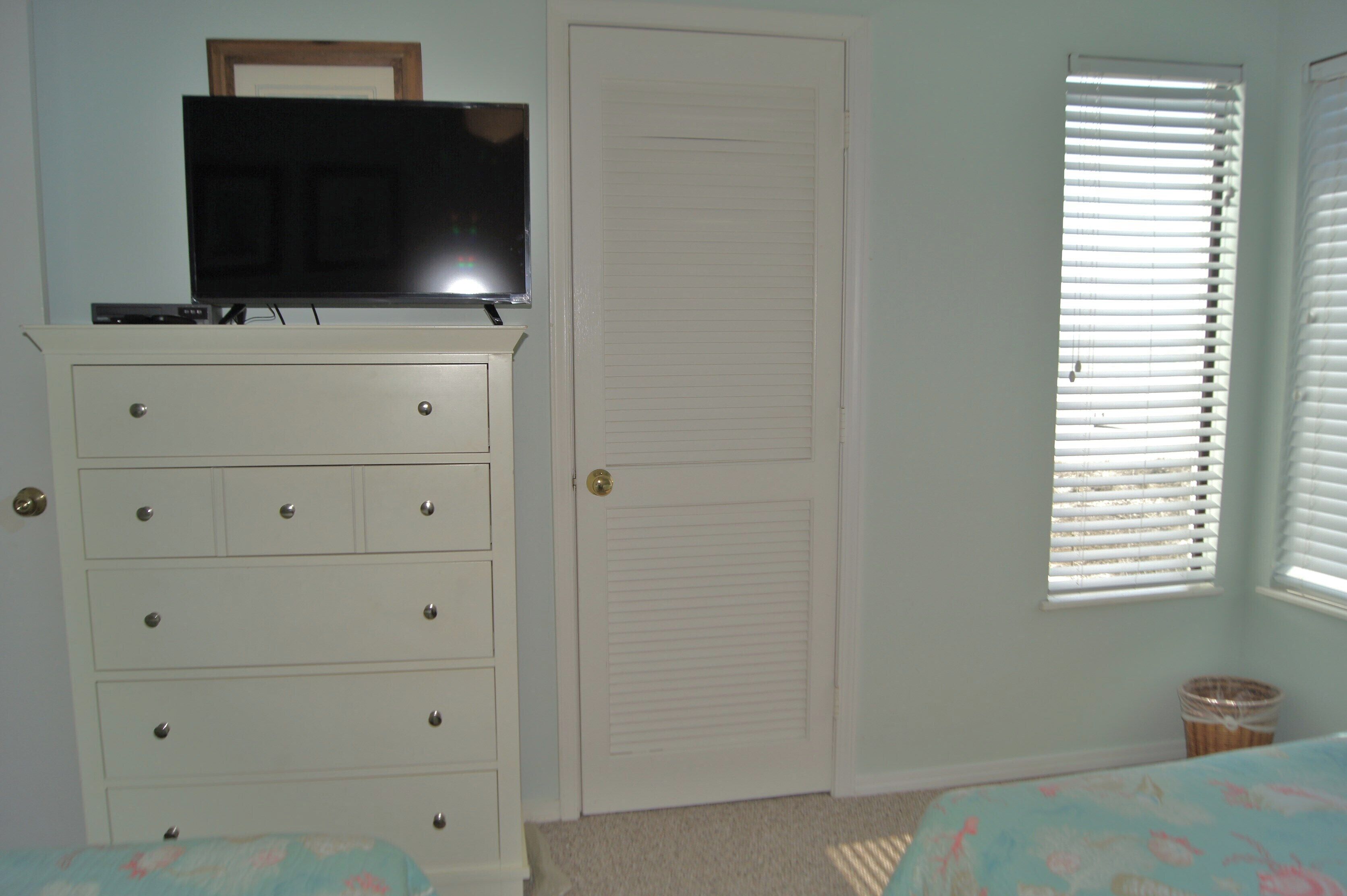 A bedroom with a dresser, a television on top, a closed door, and a window with blinds.