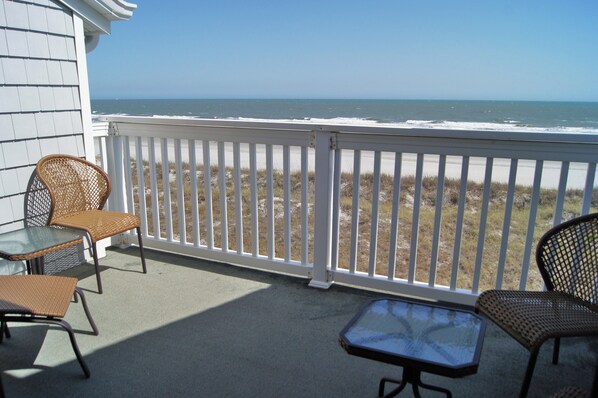 Een balkon met rieten stoelen en een tafel, met uitzicht op een strand met een witte omheining.