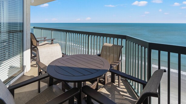 A balcony with a round table and chairs, overlooking a beach with the ocean and a clear sky. - Ashworth 1809 (North Myrtle Beach)
