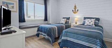Ein Schlafzimmer mit zwei Betten, einem Schreibtisch mit Fernseher und Laptop, einem Fenster mit Vorhängen und einem Deckenventilator.