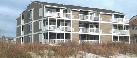 A multi-story building with balconies, located near a sandy area with vegetation.
