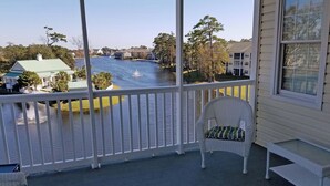 A balcony with a view of a river, a white wicker chair with a green cushion, and a small table. - Ocean Keyes 3941 (North Myrtle Beach)