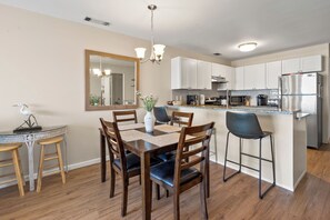 A modern kitchen with white cabinets, a stainless steel refrigerator, and a dining area with a wooden table and chairs. - Sea Cloisters 303e (North Myrtle Beach)