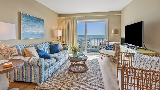 A spacious living room with a sofa, a coffee table, and a television. A balcony with a view of the ocean is visible. - Sea Cloisters 304f (North Myrtle Beach)