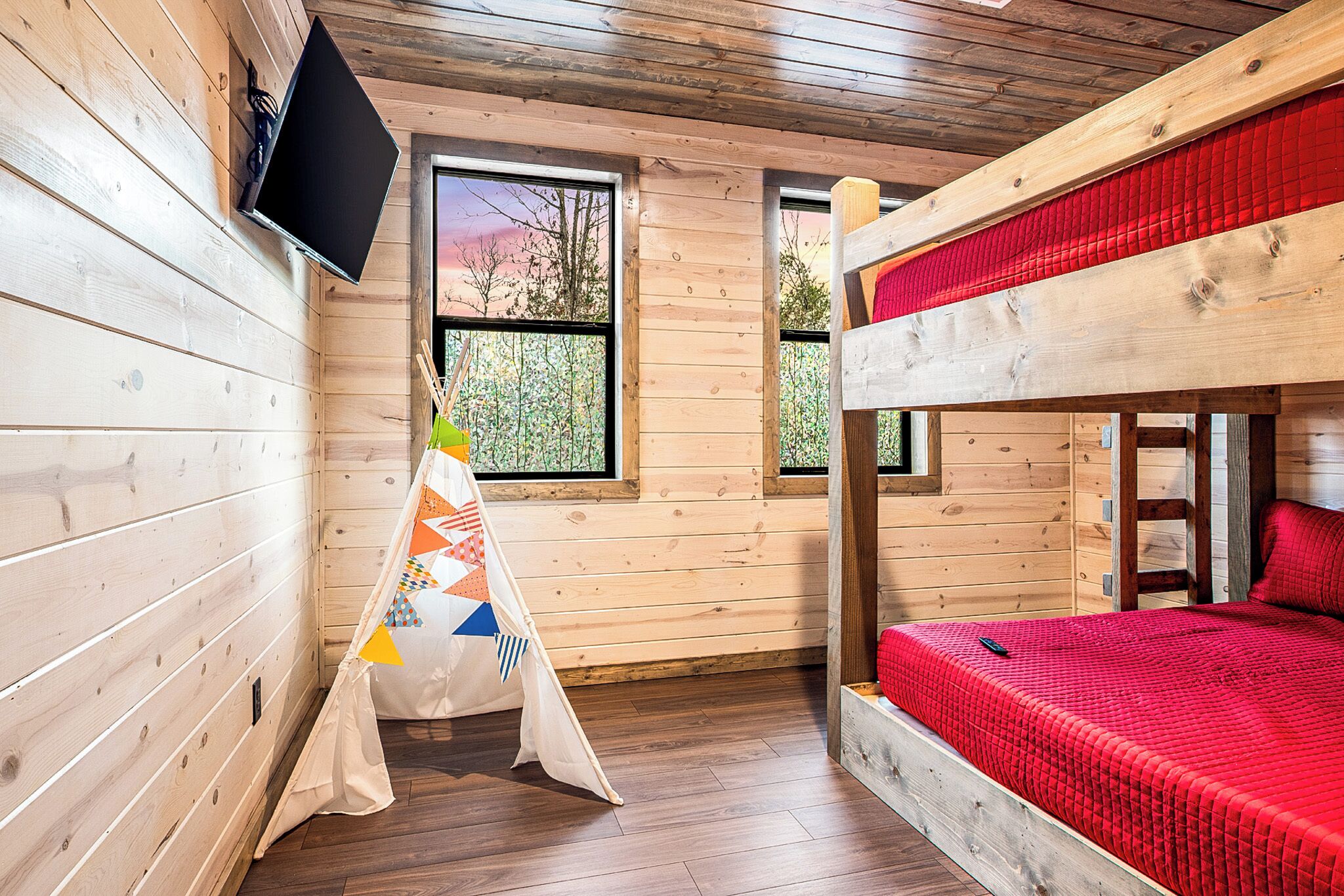 A wooden cabin room with a bunk bed, a TV mounted on the wall, and a window with a view of trees.