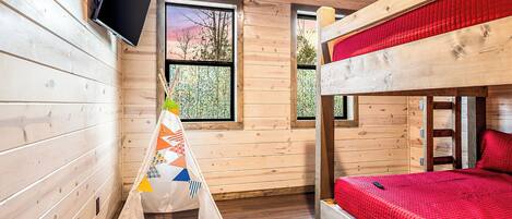 A wooden cabin room with a bunk bed, a TV mounted on the wall, and a window with a view of trees.