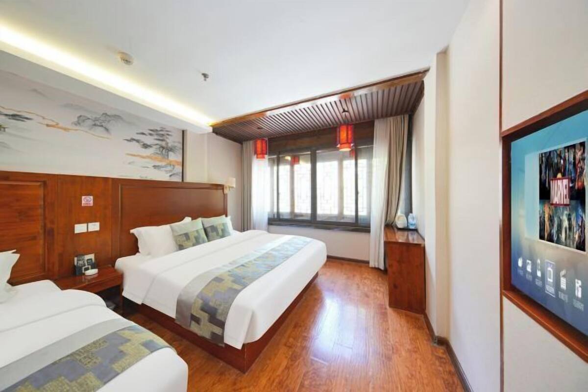 A hotel room with two beds, wooden flooring, a large window with curtains, and a wall-mounted artwork.