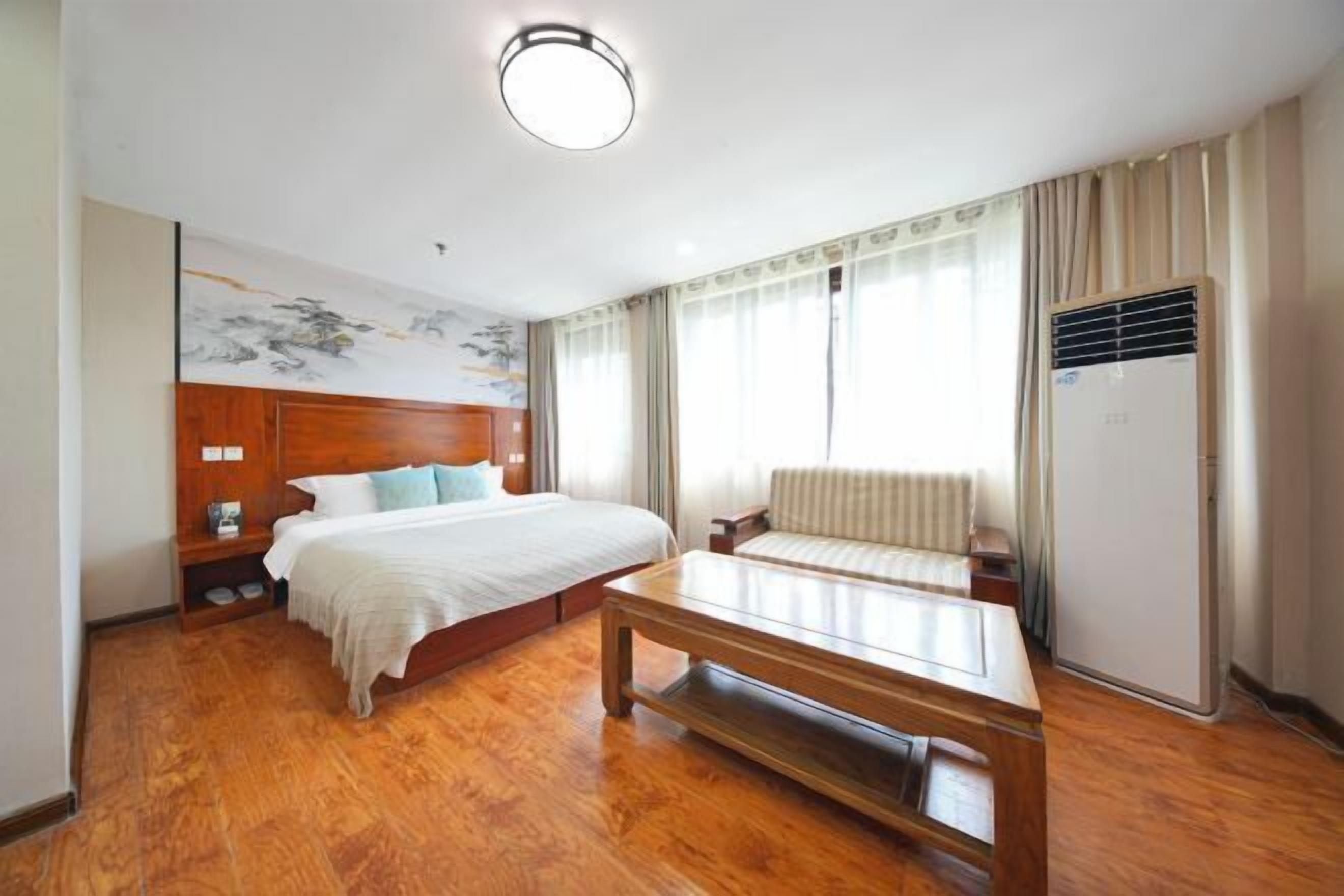 A hotel room with a wooden floor, a bed with a headboard, a bedside table, a chair, a radiator, and a window with curtains.
