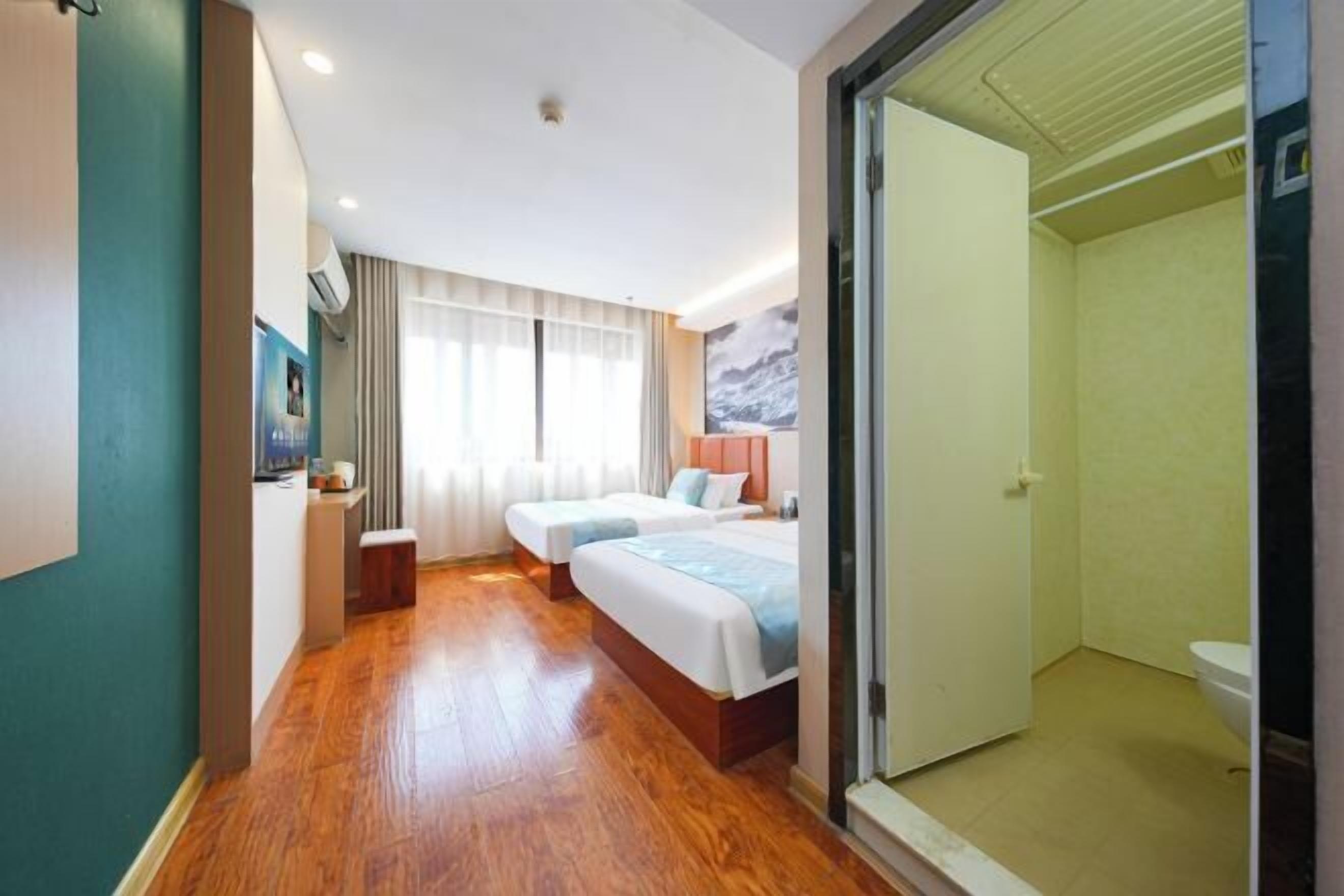 A hotel room with two beds, a wooden floor, a bathroom with a toilet, and a large window with curtains.
