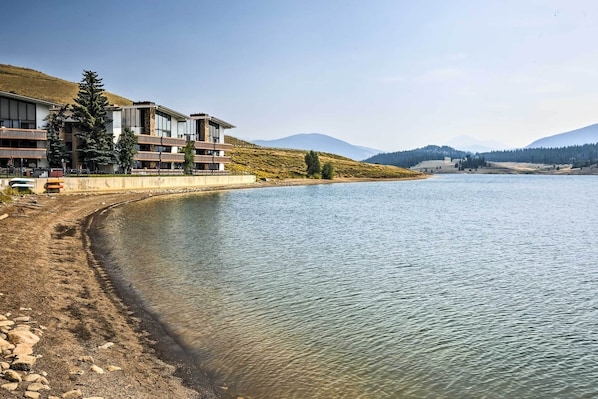 Un hotel frente al lago con varios balcones, una playa de arena y montañas al fondo.