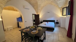 A room with a bed, a dining table with chairs, a kitchen area, and a stone ceiling.