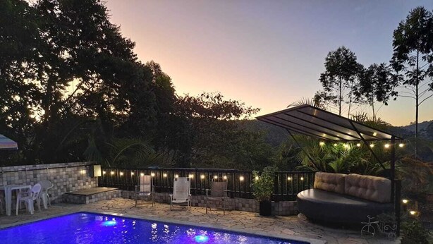 En swimmingpool med en stenkant, omgivet af udendørs siddepladser og en pergola med lyskæder.