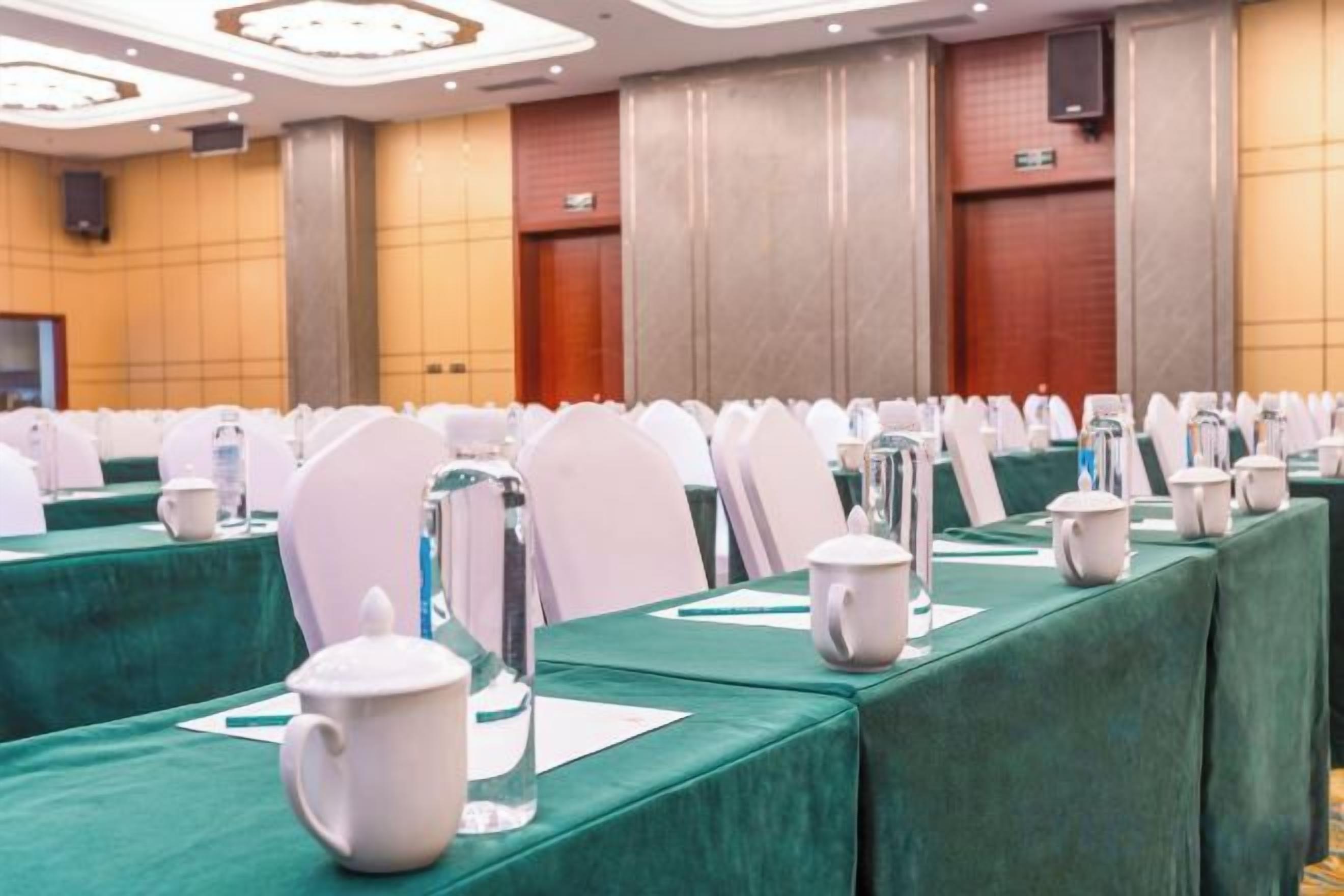 Une salle de conférence avec des rangées de tables, chacune recouverte d’une nappe verte, des chaises blanches et des carafes d’eau.