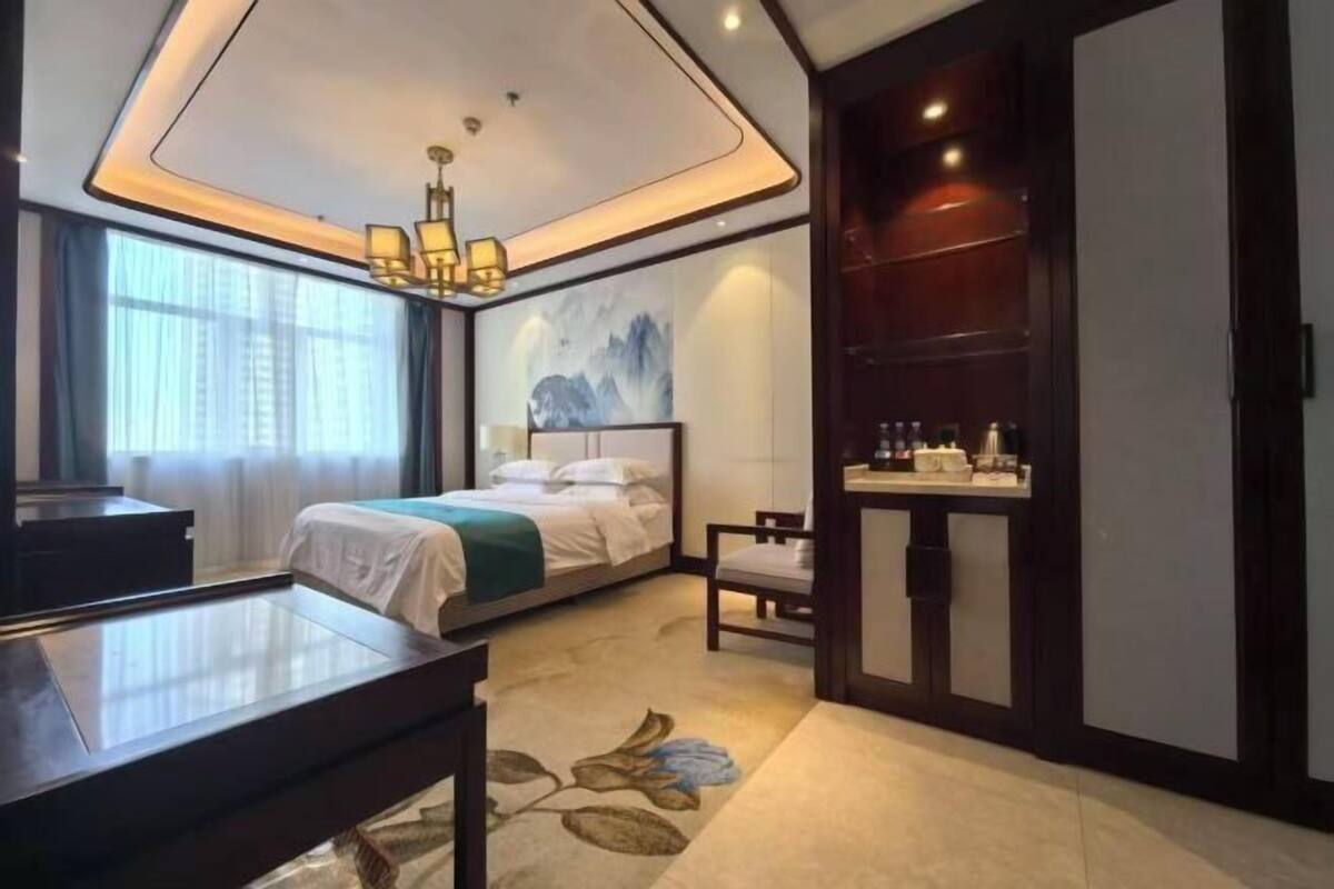 A hotel room with a bed, a desk, a chair, a small table, a painting on the wall, and a window with curtains.