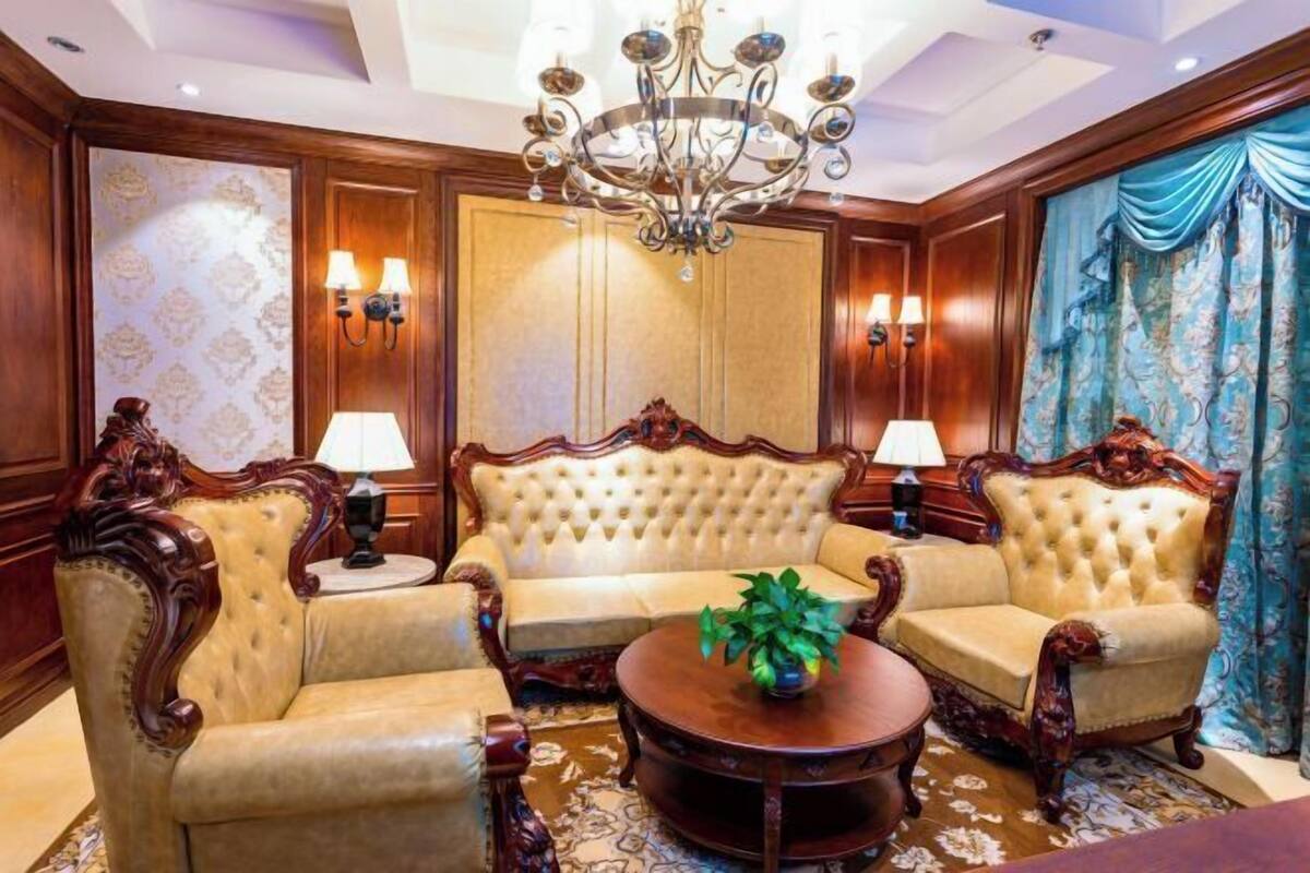 A room with a tufted sofa set, two armchairs, a round coffee table, a chandelier, wall sconces, and patterned wallpaper.