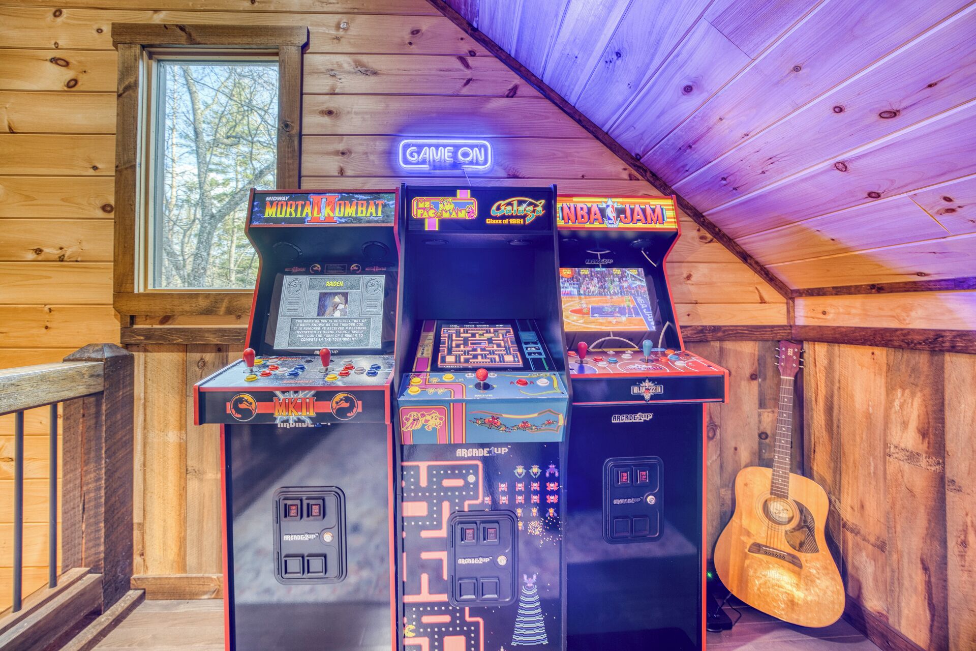 Une rangée de jeux d’arcade, dont Mortal Kombat, Galaga et NBA Jam, avec une guitare appuyée contre le mur.