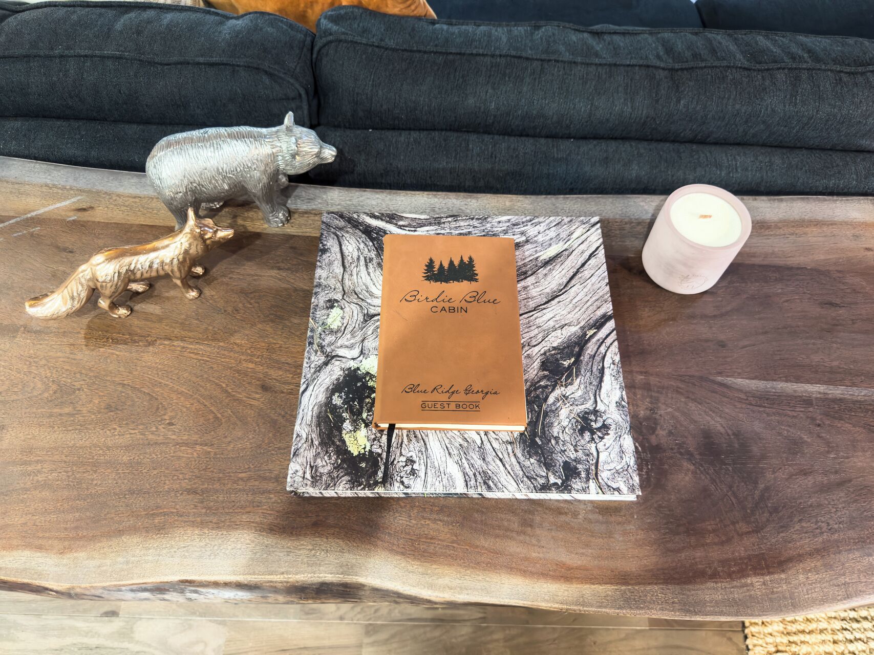 Un livre d’or intitulé « Birdie Blue Cabin » est posé sur une table en bois à motifs de marbre, à côté d’une statuette de renard décorative et d’une bougie.