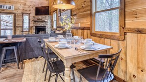 Interior de una cabaña de madera con una mesa de comedor para cuatro personas, una chimenea de piedra y un ventanal con vistas a árboles.