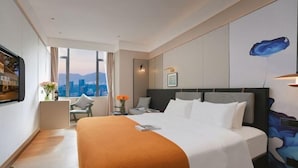 A hotel room with a bed, a desk, a chair, a window with a city view, and a wall with a painting. - Xuefu Hotel (Shenzhen Science Park Nanshan Subway Station Branch) (Shenzhen)