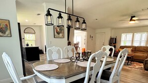 A dining area with a wooden table and white chairs, a chandelier, and a kitchen in the background.