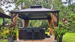 A covered outdoor seating area with a bar, surrounded by potted plants and a grassy lawn.