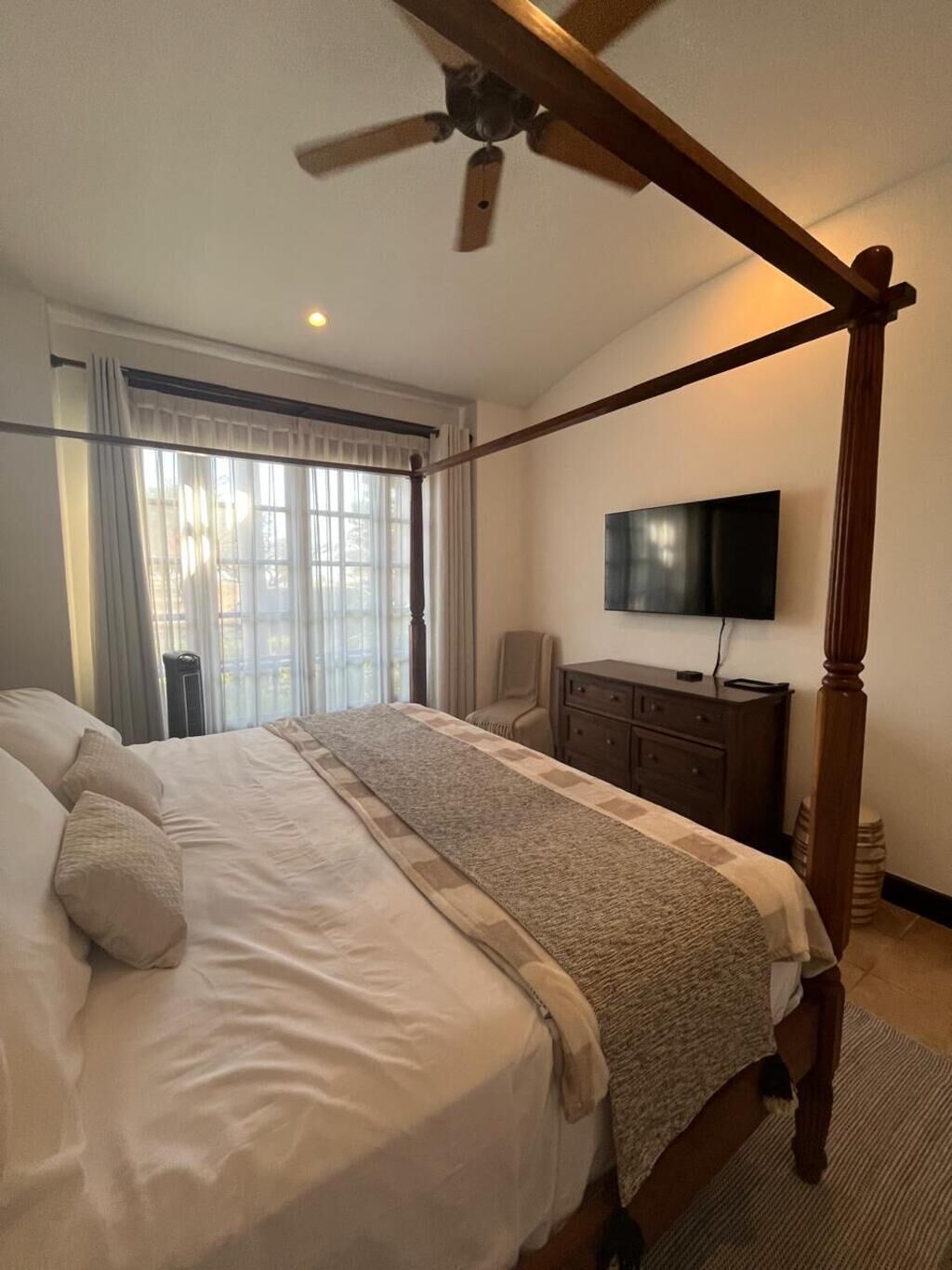 A four-poster bed with a television on the wall, a desk, and a window with curtains.