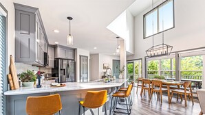 A modern kitchen with a large island, bar stools, and a dining area with wooden chairs and a table. - Lakefront | Dock, 4 Kayaks, Firepit, Sleeps 8 (Lewisburg)