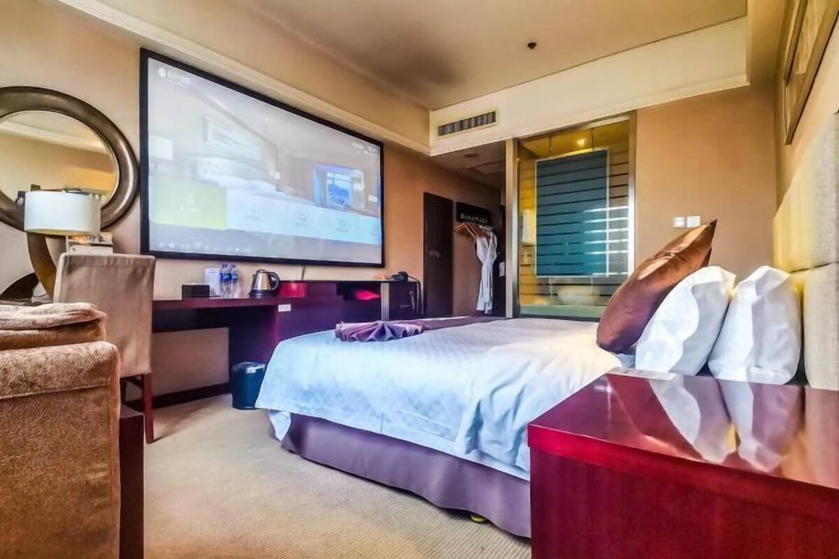 Ein Hotelzimmer mit einem großen Bett, einem Schreibtisch mit Computer, einem an der Wand montierten Fernseher und einem Fenster mit Jalousien.