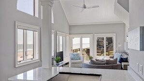 Uma cozinha com fogão, sala de jantar com mesa e cadeiras, e grandes janelas com vista para a praia.