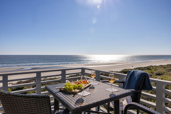 A table with a bottle, glasses, and a fruit platter on a balcony overlooking the beach.