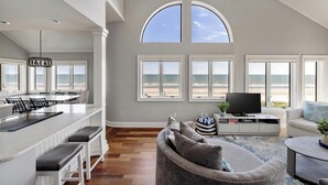 Uma sala de estar espaçosa com uma grande janela com vista para a praia, uma televisão sobre um console e uma área de estar com sofá e banquinho.