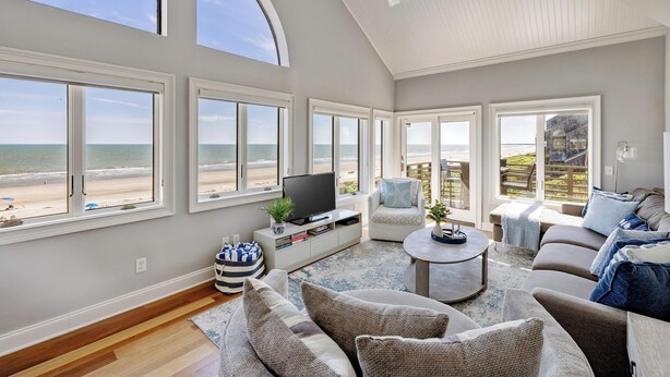 Uma sala de estar ampla com grandes janelas com vista para a praia, um ventilador de teto e uma TV de tela plana sobre um console de madeira.