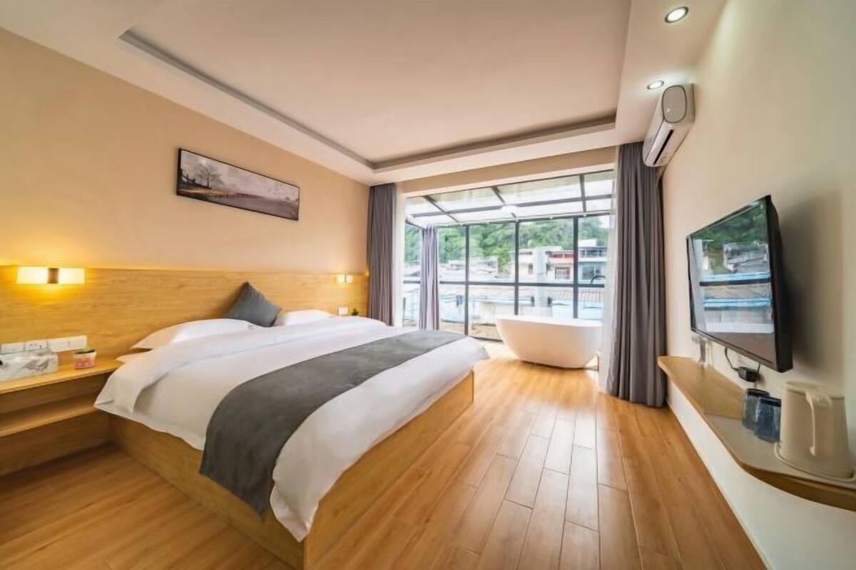 A modern hotel room with a large bed, a bathtub, a flat-screen TV, and a window with a view of the outside.