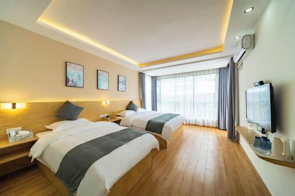 A hotel room with two beds, a flat-screen TV, a bedside table with a lamp, and a large window with curtains.