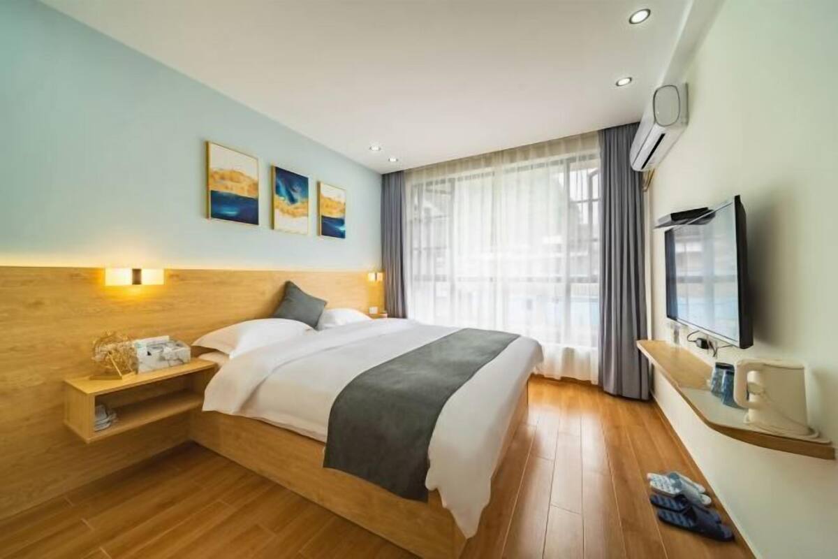 A hotel room with a wooden headboard, a bed with white and gray bedding, a bedside table with a lamp, a TV mounted on the wall, and a window with curtains.