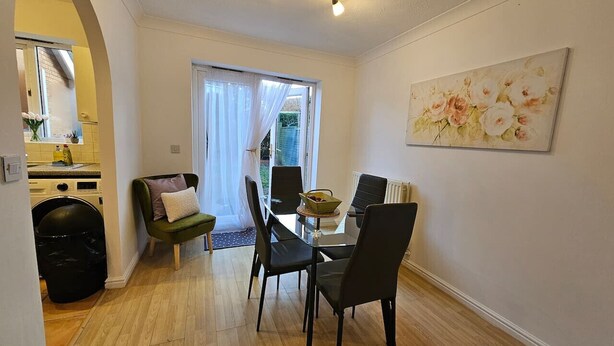 A dining area with a table and chairs, a green armchair, a black trash bin, and a window with curtains. - Sigerson House Long Stay Discounts Free Parking (Swindon)