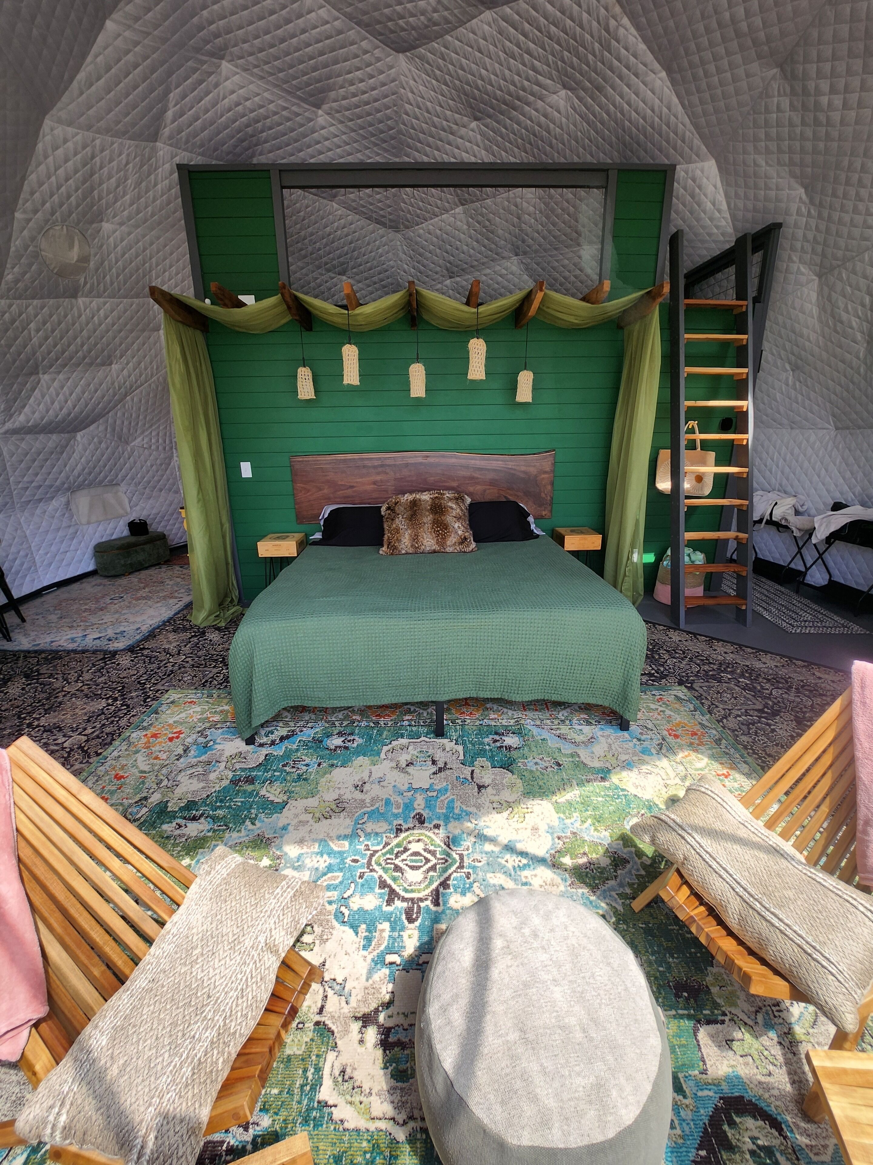 A bedroom with a green headboard, a large bed with a patterned bedspread, a wooden bench, a round ottoman, and a green wall.
