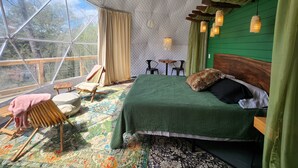 Ein gemütliches Zimmer mit einem großen Bett, einem Holzstuhl, einem kleinen Tisch und Blick durch ein großes Fenster.