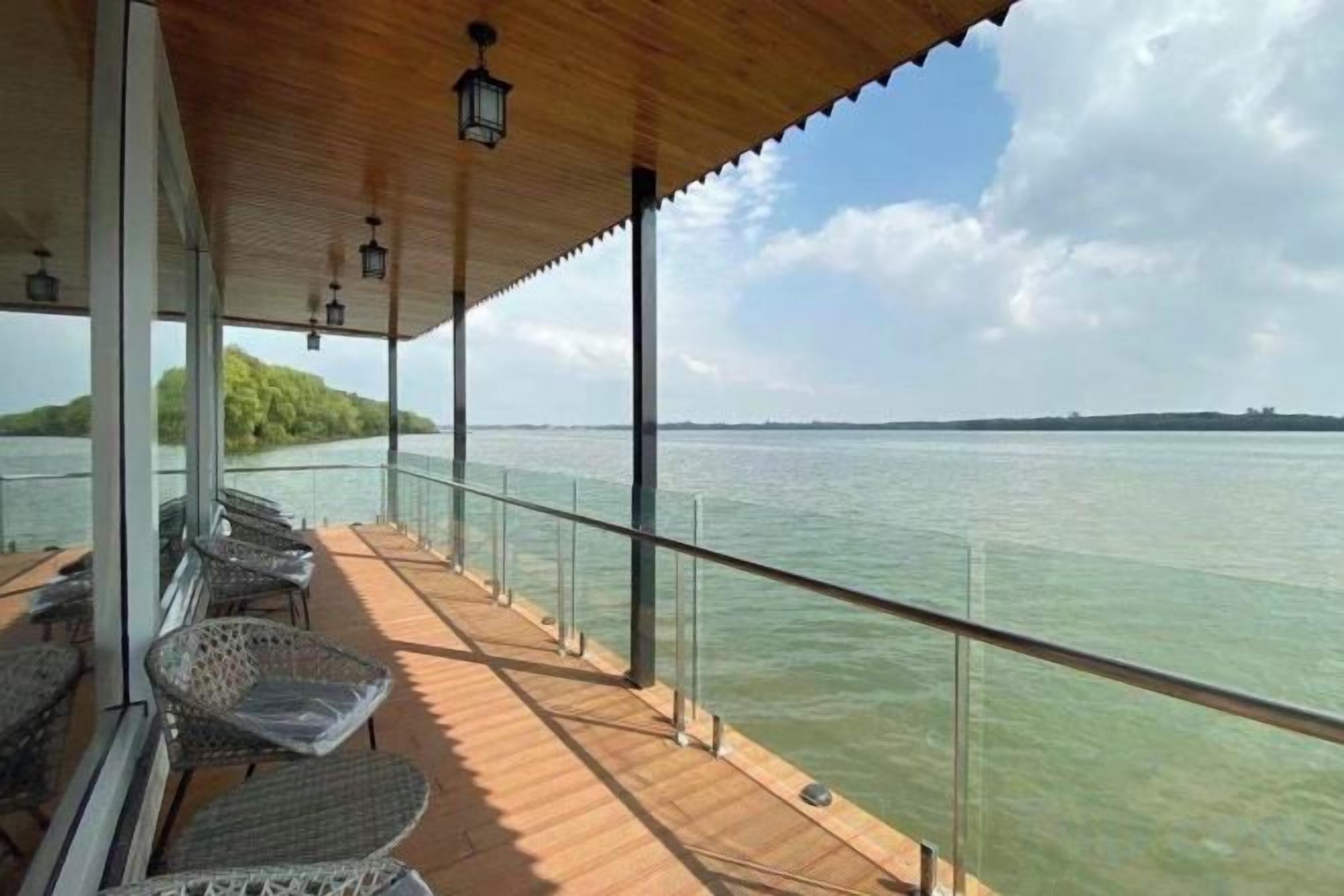 Un espacio al aire libre cubierto con vista al agua, que cuenta con una terraza de madera, barandilla de vidrio y mobiliario de mimbre.