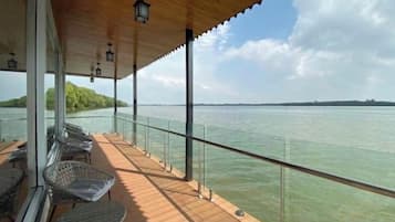 Área externa coberta com vista para a água, com deck de madeira, guarda-corpo de vidro e móveis de vime.