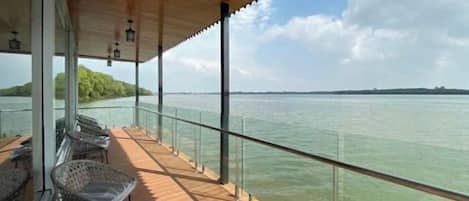 Ein überdachter Außenbereich mit Blick auf das Wasser, ausgestattet mit einem Holzboden, einer Glasumlaufung und Korbmöbeln.
