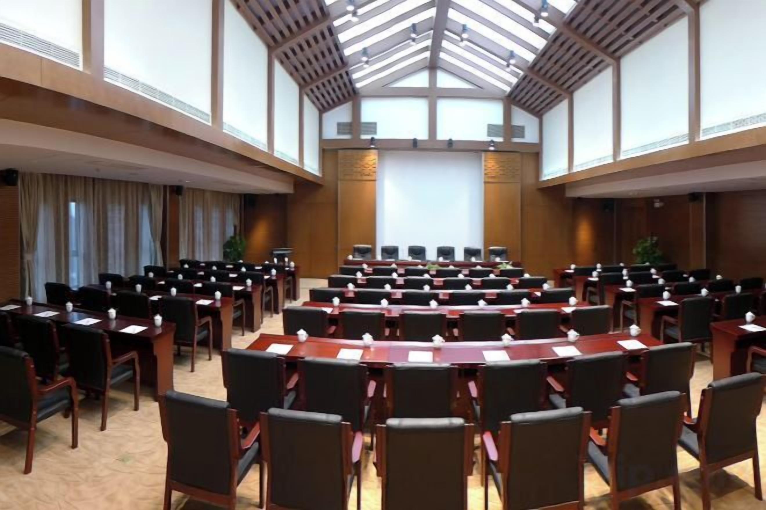 Ein Konferenzraum mit Reihen von Tischen und Stühlen, einer großen Leinwand vorne und einer hohen Decke mit Oberlichtern.