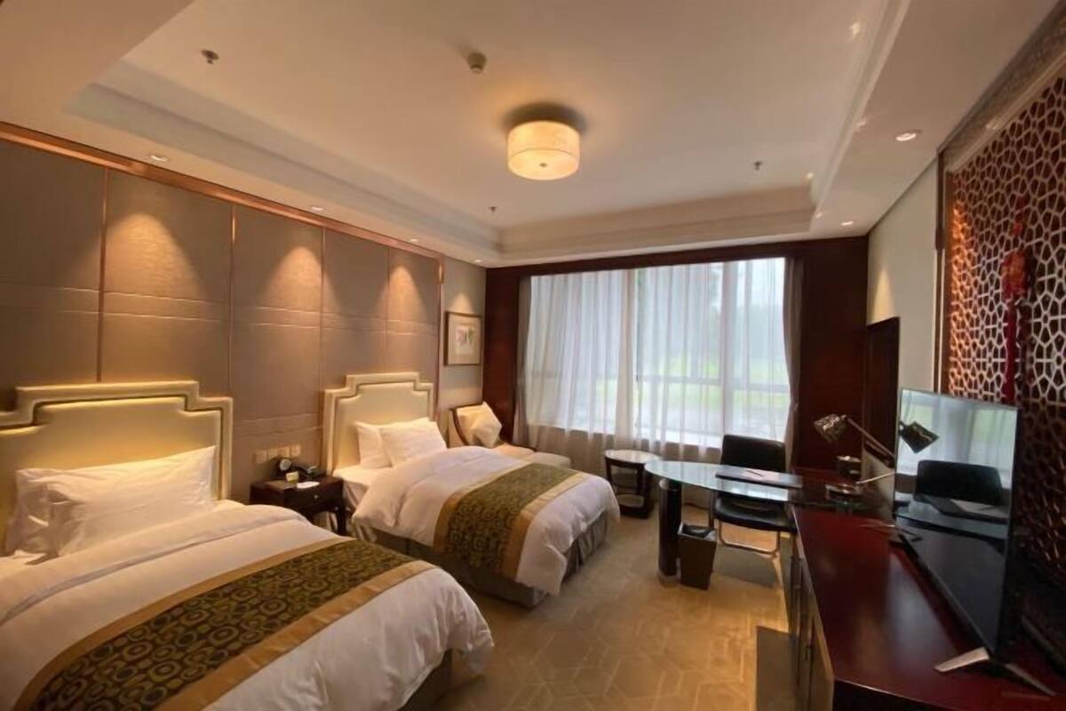 A hotel room with two beds, a desk, a chair, a TV, and a large window with curtains.