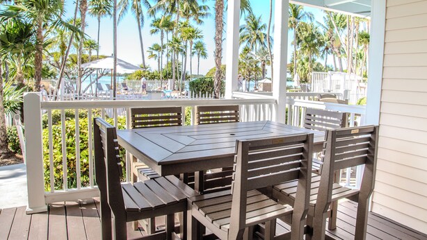 A wooden deck with a table and chairs, surrounded by a white fence, overlooking a pool area with palm trees and lounge chairs.