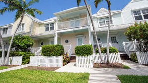 A row of two-story houses with a white picket fence, a front porch, and a small front yard with palm trees and shrubs.