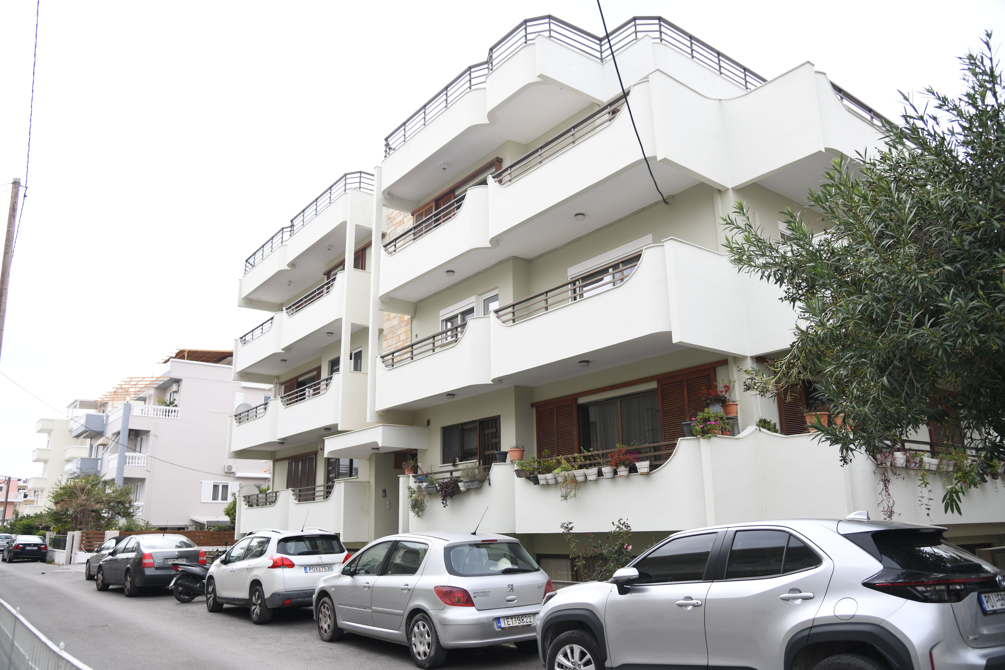 Un immeuble résidentiel à plusieurs étages, doté de balcons, avec des voitures garées et des plantes en pot.