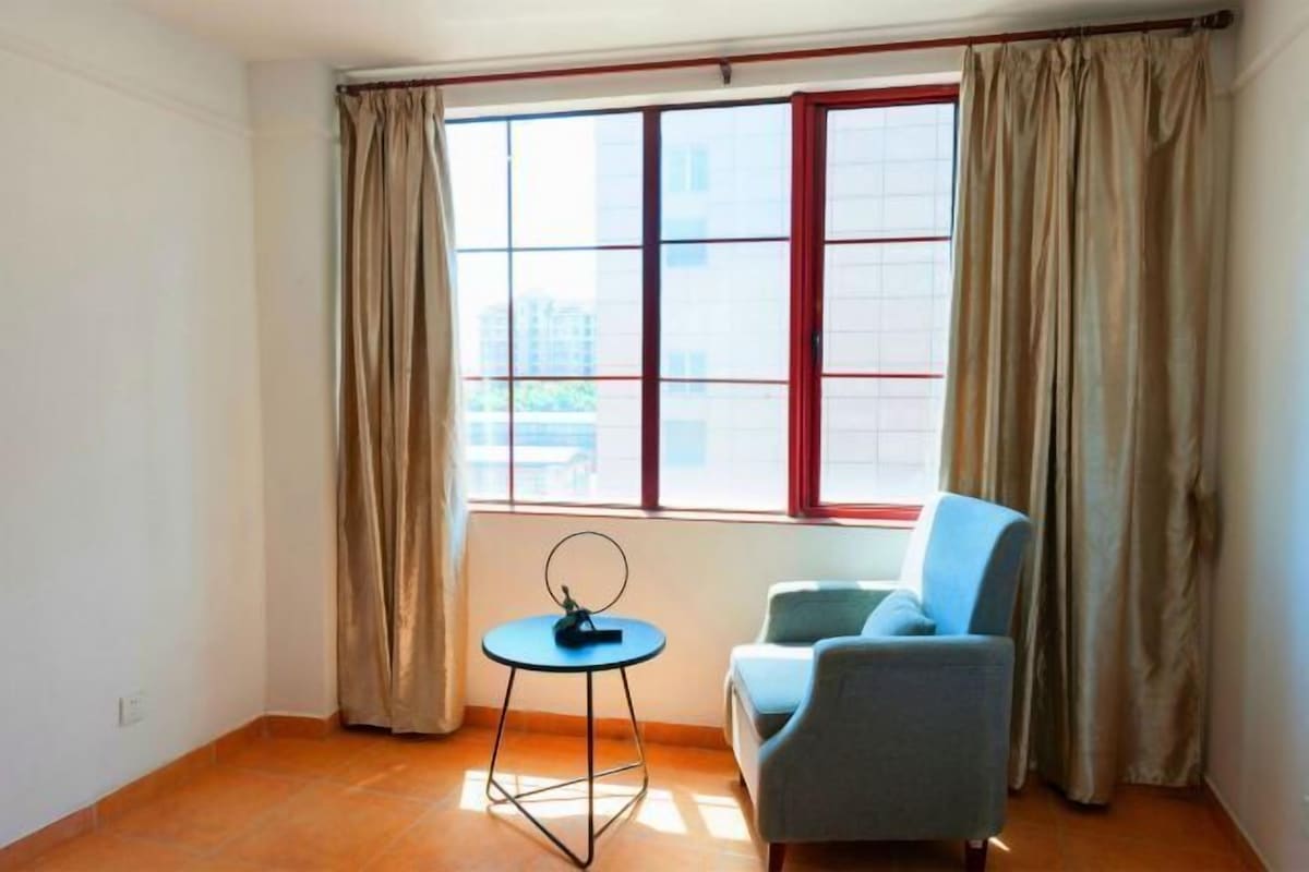 A room with a single blue chair, a small round table, a window with curtains, and a tiled floor.