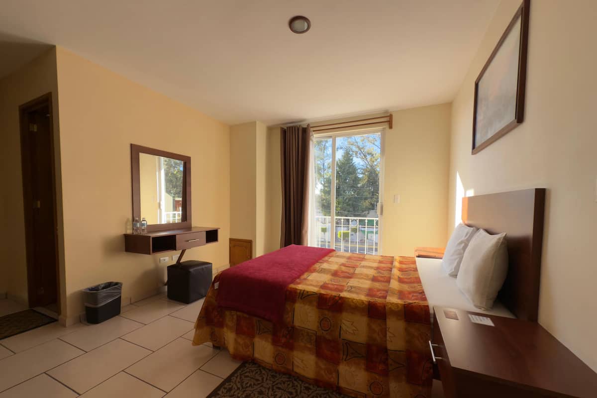 A hotel room with a bed, a bedside table, a mirror, a window, and a view of the outdoors.