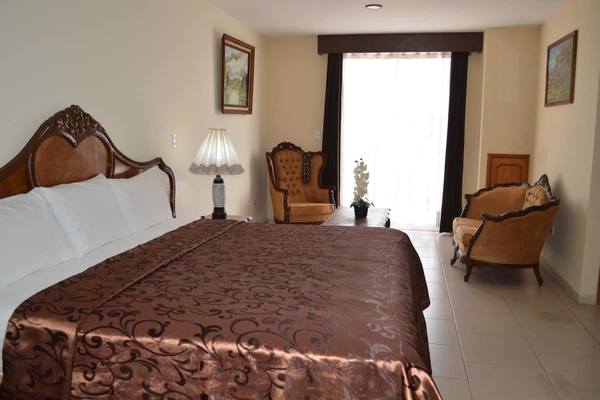 A hotel room with a large bed, two armchairs, a lamp, and a potted plant.
